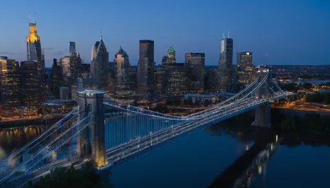 A lit suspension bridge crosses a dark river with a glowing city skyline in the background.