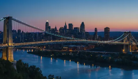 A lit suspension bridge crosses a river with a city skyline glowing at dusk.