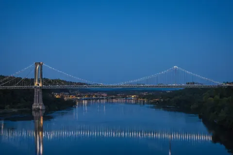 A lit suspension bridge reflects in calm water under a twilight sky