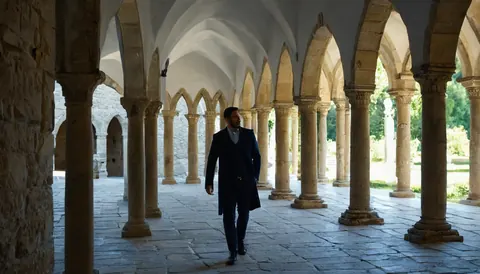 A man in a dark suit walks through a sunlit stone cloister with repeating arches and columns.