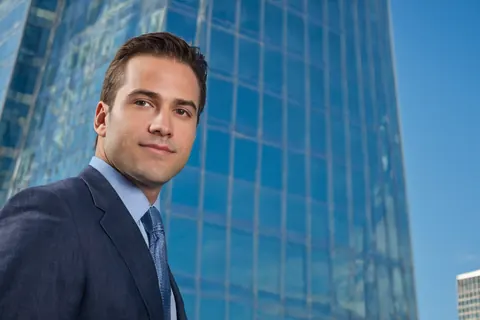 A man in a suit and tie stands in front of a modern glass skyscraper on a clear day.