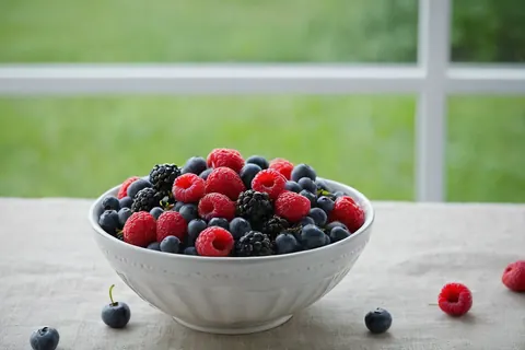 A white bowl filled with mixed berries on a linen tablecloth near a window
