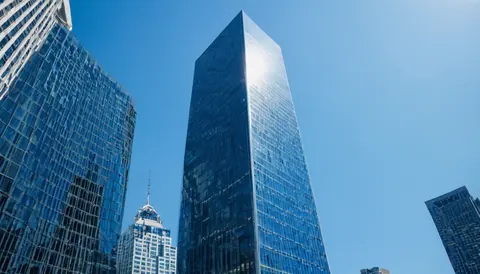 Modern glass skyscrapers rise against a clear blue sky with sunlight reflecting off their surfaces.