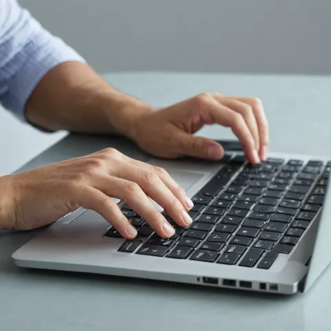 Person typing on a silver laptop keyboard with black keys on a gray desk.