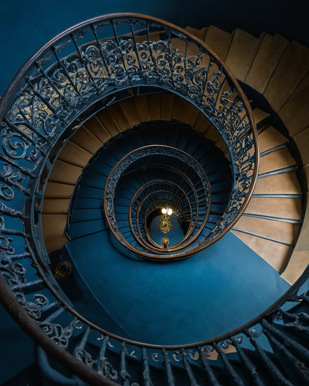 Spiral staircase with ornate iron railings and wooden steps descending into blue walls