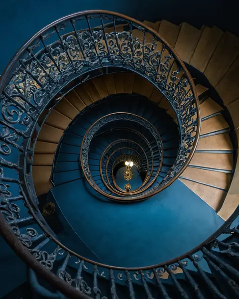 Spiral staircase with ornate iron railings and wooden steps descending into blue walls